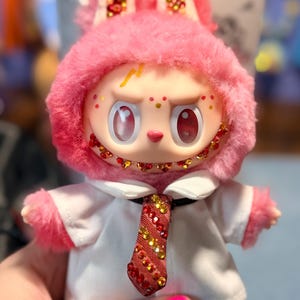 May include: A plush doll with pink fur, wearing a white collared shirt and a red and gold striped tie. The doll features large red eyes, a wide grin with red and gold rhinestones, and matching rhinestones on its ears and tie. A yellow lightning bolt is painted on its forehead.