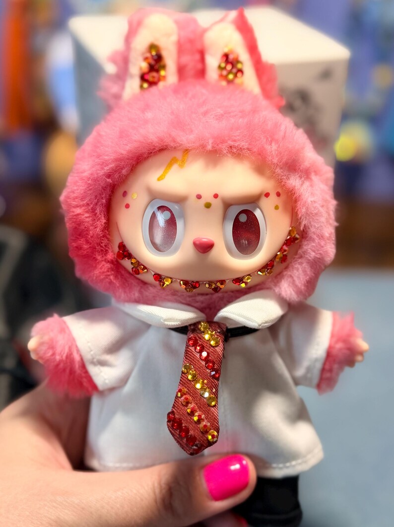 May include: A plush doll with pink fur, wearing a white collared shirt and a red and gold striped tie. The doll features large red eyes, a wide grin with red and gold rhinestones, and matching rhinestones on its ears and tie. A yellow lightning bolt is painted on its forehead.