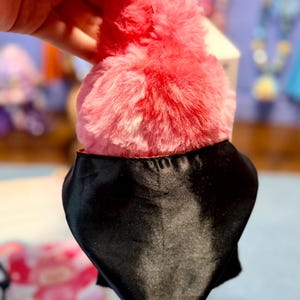 May include: A plush toy featuring a vibrant pink and red fuzzy texture on the top portion, contrasted by a smooth, solid black satin-like fabric covering the lower section. The object has a rounded, soft appearance with a distinct color transition.
