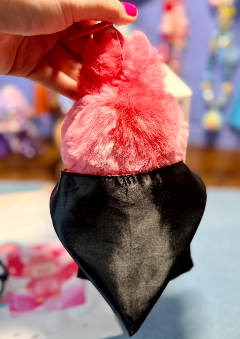 May include: A plush toy featuring a vibrant pink and red fuzzy texture on the top portion, contrasted by a smooth, solid black satin-like fabric covering the lower section. The object has a rounded, soft appearance with a distinct color transition.