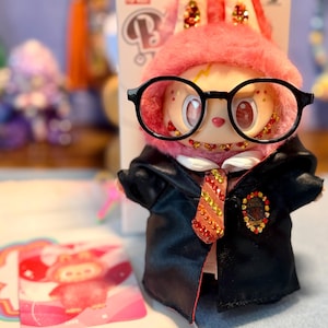 May include: A collectible vinyl figure wearing a pink fuzzy hood with sequined ears, large black-rimmed glasses, and a black satin robe over a white shirt. The figure features a red and gold sequined tie and a Magic School crest, with a matching character card.