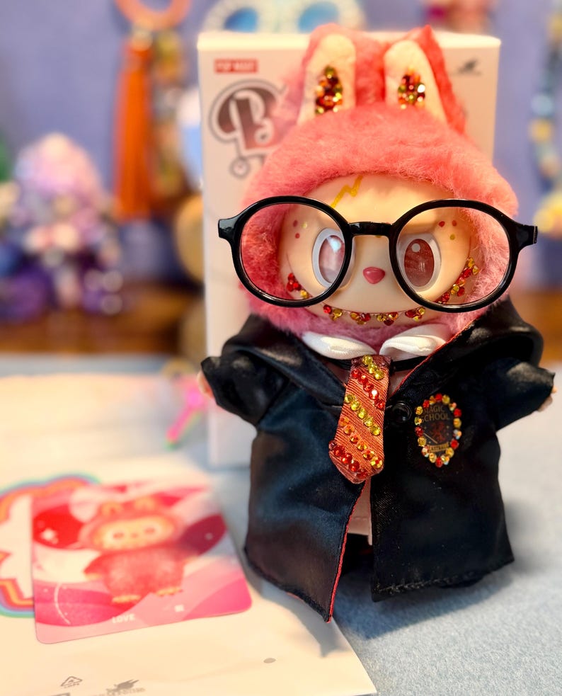 May include: A collectible vinyl figure wearing a pink fuzzy hood with sequined ears, large black-rimmed glasses, and a black satin robe over a white shirt. The figure features a red and gold sequined tie and a Magic School crest, with a matching character card.