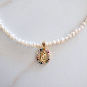 Pu&ograve; includere: Collana di perle bianche irregolari con un ciondolo dorato raffigurante una figura religiosa. Il ciondolo &egrave; circondato da un'aureola di pietre preziose colorate. Un gioiello delicato.