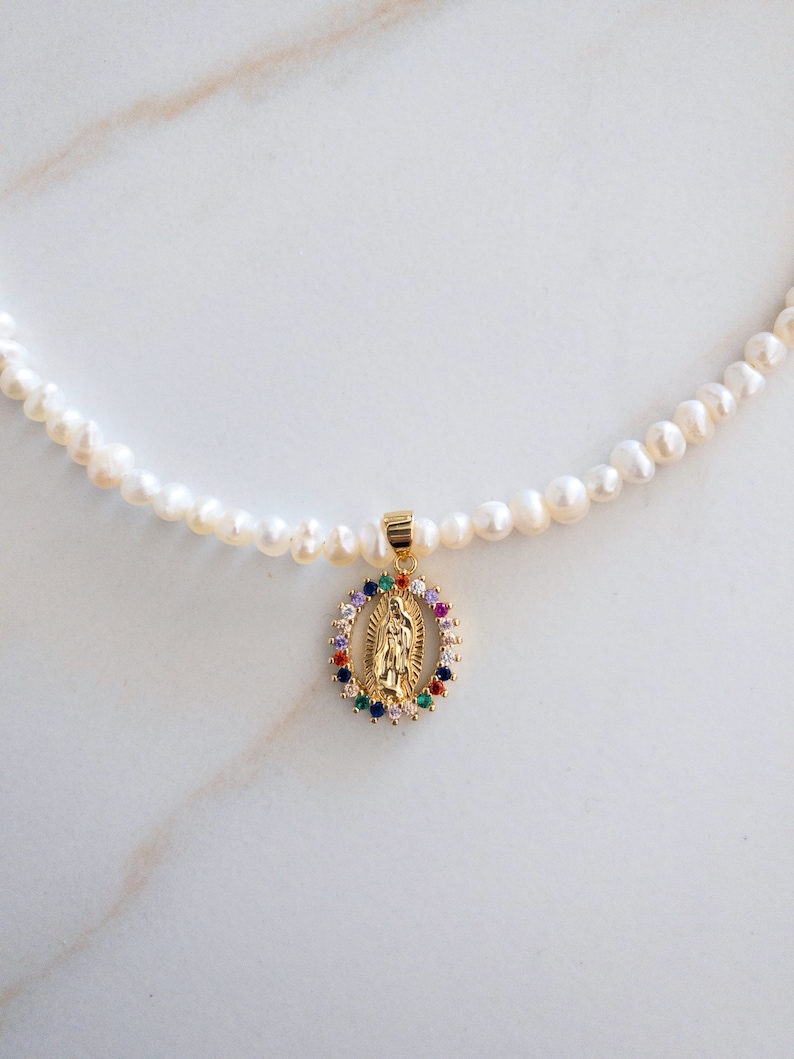 Pu&ograve; includere: Collana di perle bianche irregolari con un ciondolo dorato raffigurante una figura religiosa. Il ciondolo &egrave; circondato da un'aureola di pietre preziose colorate. Un gioiello delicato.
