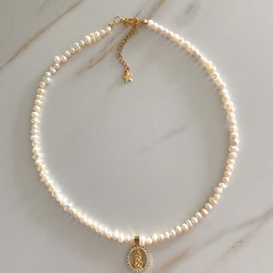 Pu&ograve; includere: Una delicata collana di perle con un ciondolo dorato raffigurante una figura religiosa circondata da piccole pietre trasparenti. La collana ha una chiusura dorata e un piccolo ciondolo a forma di stella. Le perle sono piccole e bianche.