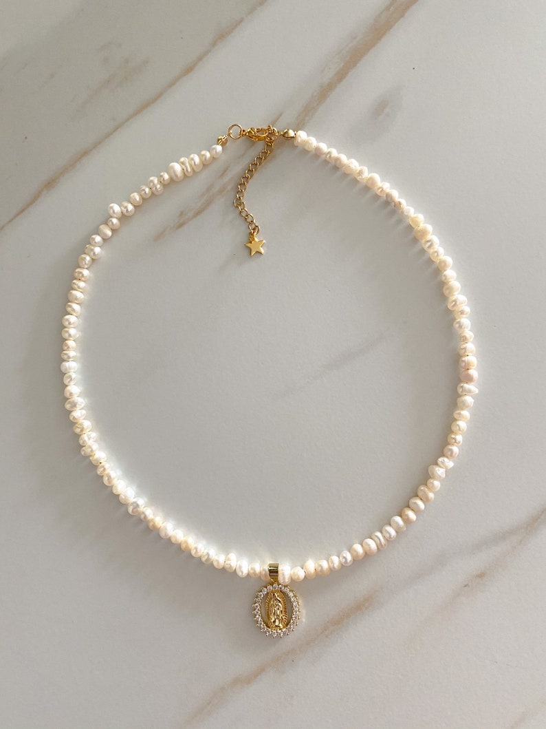 Pu&ograve; includere: Una delicata collana di perle con un ciondolo dorato raffigurante una figura religiosa circondata da piccole pietre trasparenti. La collana ha una chiusura dorata e un piccolo ciondolo a forma di stella. Le perle sono piccole e bianche.