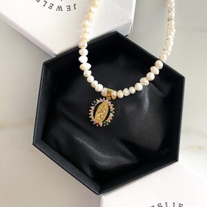Pu&ograve; includere: Una collana di perle con un ciondolo d'oro raffigurante una figura religiosa, presentata in una scatola esagonale nera. Il ciondolo &egrave; adornato con pietre preziose colorate. La collana &egrave; su una superficie bianca, con una scatola che dice "JEWELRY BY" sullo sfondo.