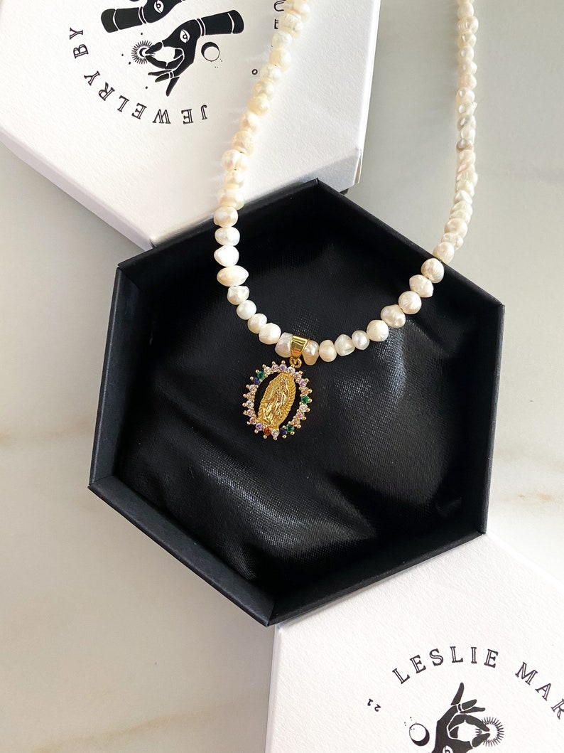 Pu&ograve; includere: Una collana di perle con un ciondolo d'oro raffigurante una figura religiosa, presentata in una scatola esagonale nera. Il ciondolo &egrave; adornato con pietre preziose colorate. La collana &egrave; su una superficie bianca, con una scatola che dice "JEWELRY BY" sullo sfondo.