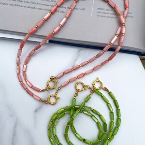 May include: Two necklaces made of shell beads. One necklace is made of pink shell beads and the other is made of green shell beads. Both necklaces have gold clasps.