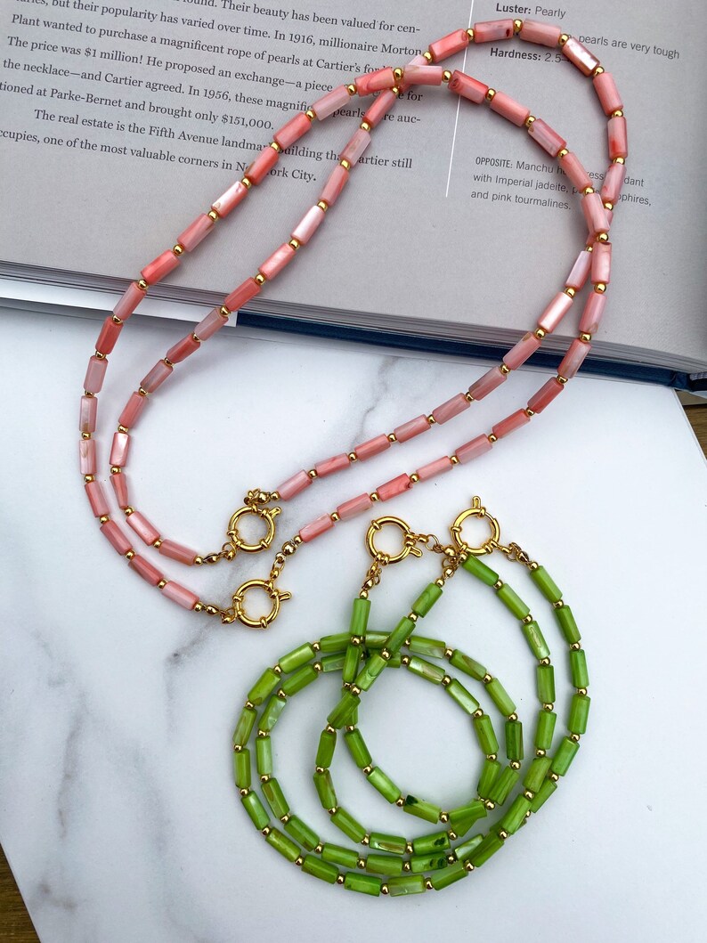 May include: Two necklaces made of shell beads. One necklace is made of pink shell beads and the other is made of green shell beads. Both necklaces have gold clasps.
