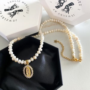 Pu&ograve; includere: Una collana di perle con un ciondolo dorato raffigurante una figura religiosa, presentata in una scatola nera. La collana &egrave; accompagnata da due scatole bianche con la scritta "LESLIE MARQUEZ JEWELRY by". Il ciondolo &egrave; circondato da piccole pietre trasparenti.