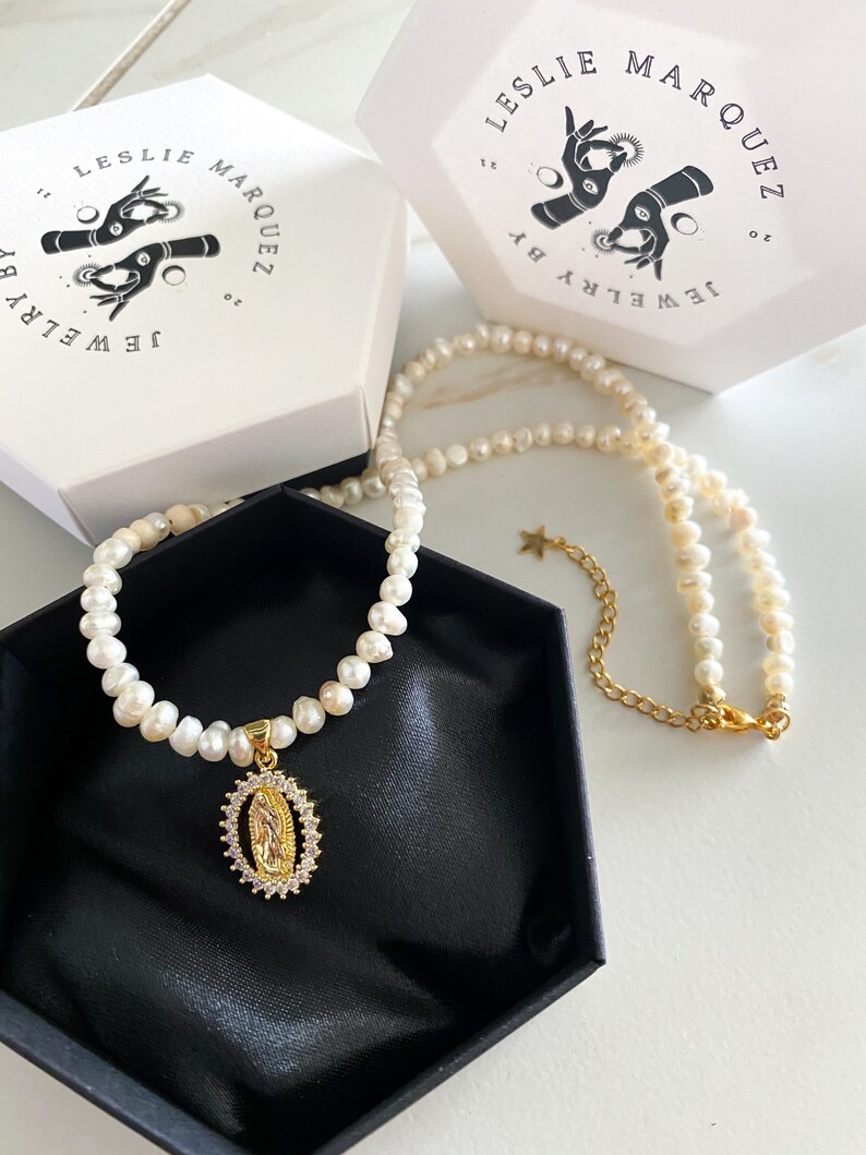 Pu&ograve; includere: Una collana di perle con un ciondolo dorato raffigurante una figura religiosa, presentata in una scatola nera. La collana &egrave; accompagnata da due scatole bianche con la scritta "LESLIE MARQUEZ JEWELRY by". Il ciondolo &egrave; circondato da piccole pietre trasparenti.