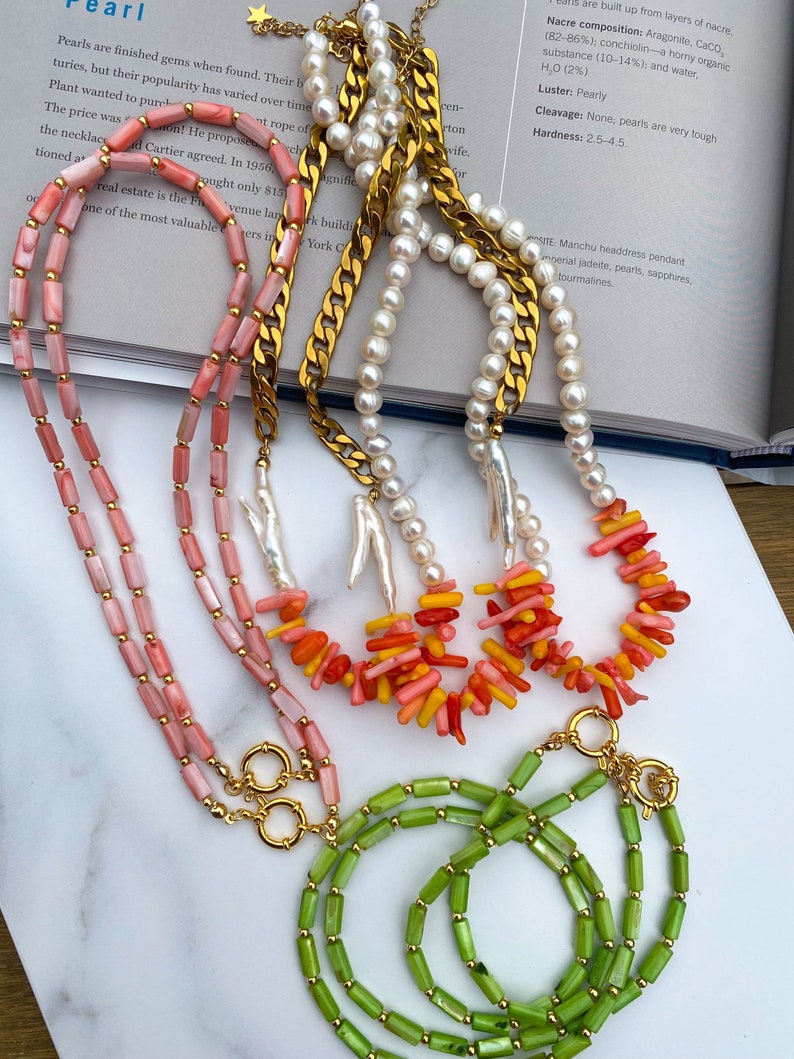 May include: Three necklaces with gold clasps. The top necklace is made of white pearls. The middle necklace is made of pink and orange coral. The bottom necklace is made of green jade.