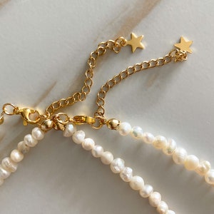 Pu&ograve; includere: Una delicata collana di perle con catena dorata e dettagli a forma di stella. La collana presenta piccole perle bianche di forma irregolare e una chiusura dorata. La catena &egrave; regolabile.