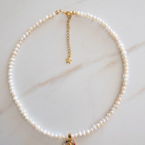Pu&ograve; includere: Una delicata collana con una fila di piccole perle bianche. Un ciondolo dorato con un alone di pietre preziose colorate e una figura religiosa pende al centro. La collana ha una catena e una chiusura dorate.