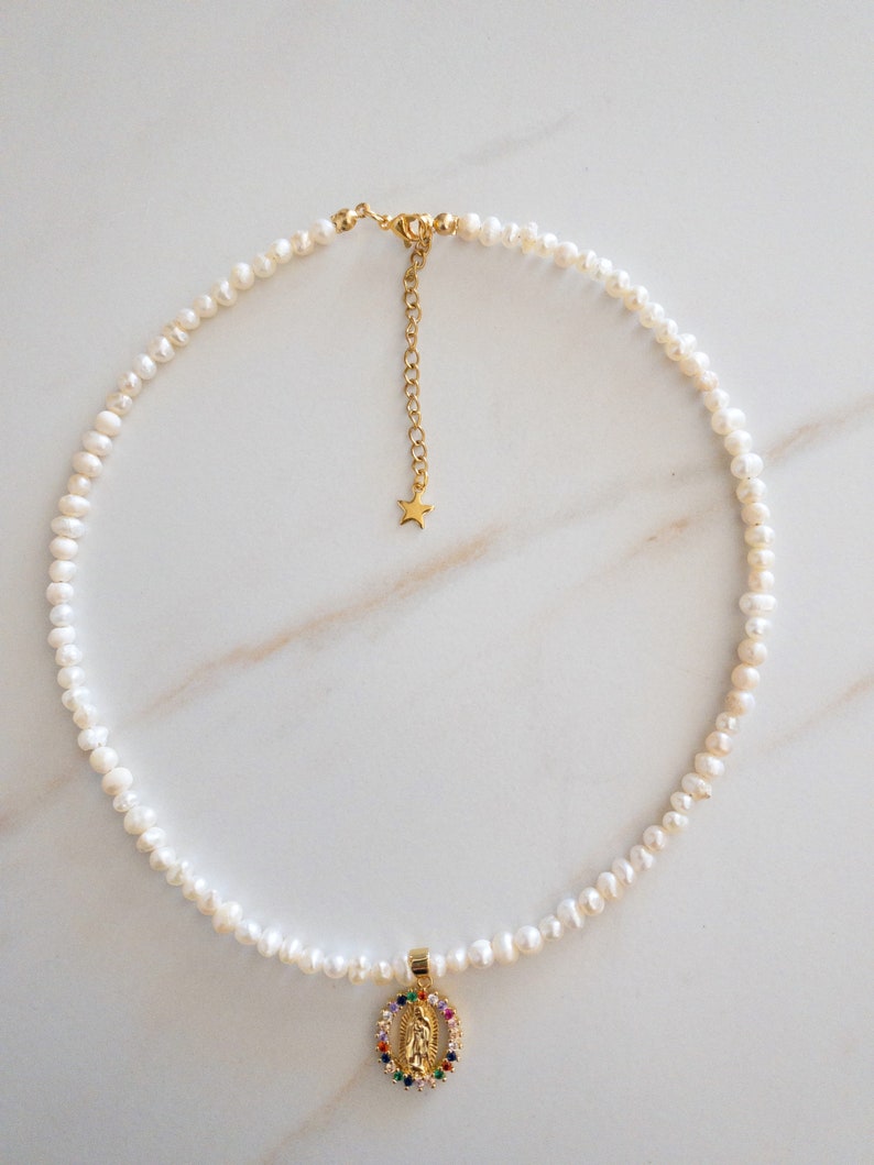Pu&ograve; includere: Una delicata collana con una fila di piccole perle bianche. Un ciondolo dorato con un alone di pietre preziose colorate e una figura religiosa pende al centro. La collana ha una catena e una chiusura dorate.