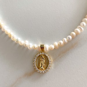 Pu&ograve; includere: Una delicata collana con una fila di perle bianche. Un ciondolo dorato con una figura religiosa &egrave; appeso alla collana. Il ciondolo &egrave; circondato da un alone di piccoli cristalli trasparenti. La collana &egrave; esposta su una superficie bianca.