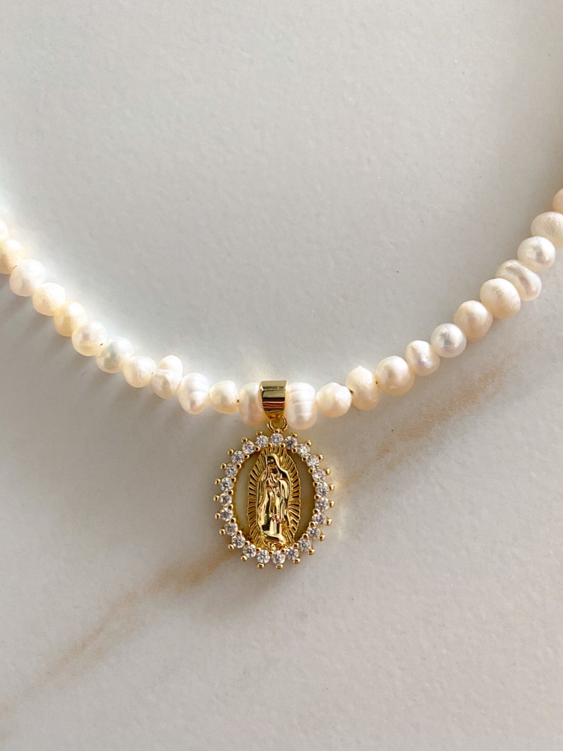 Pu&ograve; includere: Una delicata collana con una fila di perle bianche. Un ciondolo dorato con una figura religiosa &egrave; appeso alla collana. Il ciondolo &egrave; circondato da un alone di piccoli cristalli trasparenti. La collana &egrave; esposta su una superficie bianca.