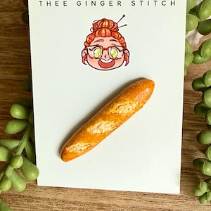 May include: A miniature baguette pin attached to a white display card featuring the text THEE GINGER STITCH and a cartoon illustration of a person with orange hair in a bun wearing glasses.