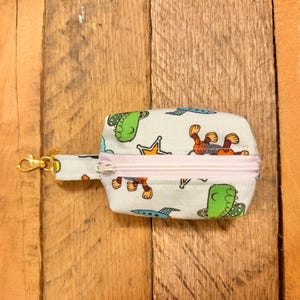 May include: A small, rectangular pouch with a light blue background and a repeating pattern of cartoon dinosaurs, monkeys, crowns, and spaceships. The pouch has a pink zipper and a gold-colored clasp.