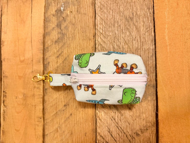 May include: A small, rectangular pouch with a light blue background and a repeating pattern of cartoon dinosaurs, monkeys, crowns, and spaceships. The pouch has a pink zipper and a gold-colored clasp.