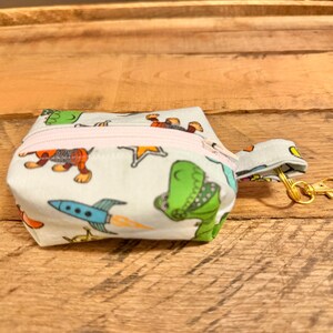 May include: A small, white fabric pouch with a light pink zipper, featuring a colorful dinosaur and rocket ship pattern. It has a small loop and a gold-colored clasp. The pouch is set against a wooden background.