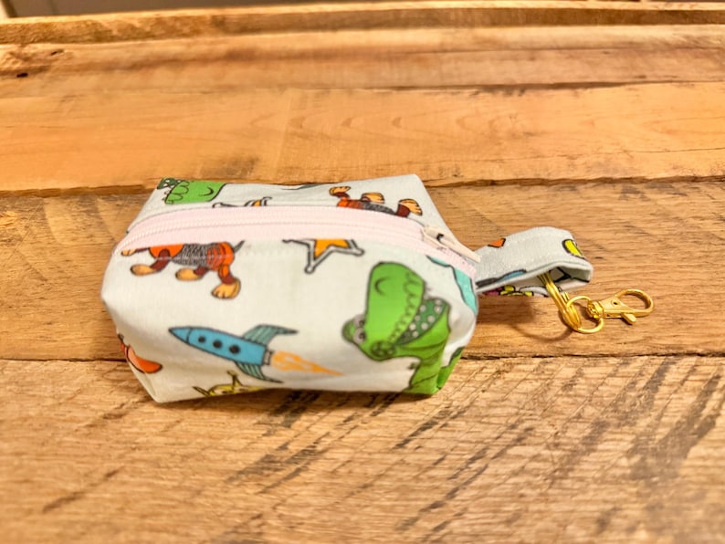 May include: A small, white fabric pouch with a light pink zipper, featuring a colorful dinosaur and rocket ship pattern. It has a small loop and a gold-colored clasp. The pouch is set against a wooden background.