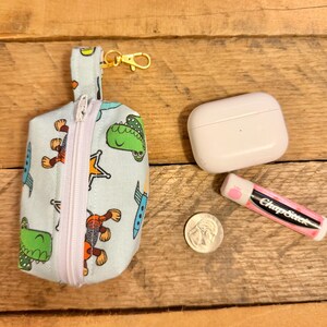 May include: A light blue fabric pouch with a zipper, a gold-colored clasp, and a small loop. The pouch is decorated with cartoon dinosaurs, rockets, and stars. Next to the pouch are a white case, a ChapStick, and a quarter.