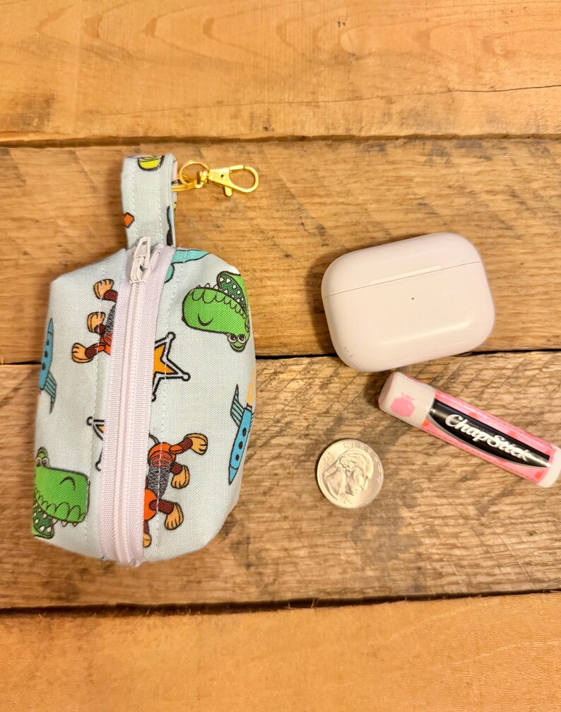 May include: A light blue fabric pouch with a zipper, a gold-colored clasp, and a small loop. The pouch is decorated with cartoon dinosaurs, rockets, and stars. Next to the pouch are a white case, a ChapStick, and a quarter.