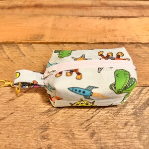 May include: A small, light blue fabric pouch with a zipper closure and a gold-colored clasp. The pouch is decorated with cartoon characters, including a green dinosaur, a rocket ship, and a three-eyed alien. The zipper is light pink.