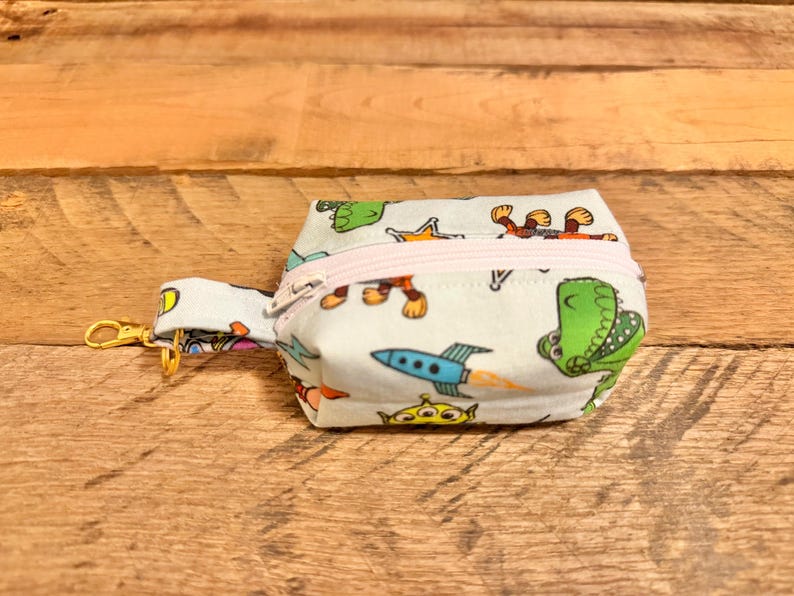 May include: A small, light blue fabric pouch with a zipper closure and a gold-colored clasp. The pouch is decorated with cartoon characters, including a green dinosaur, a rocket ship, and a three-eyed alien. The zipper is light pink.