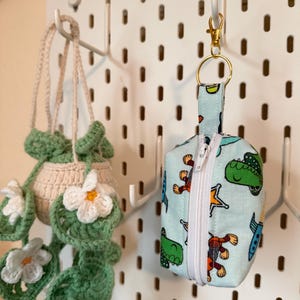 May include: A light blue zippered pouch with a cartoon dinosaur print, attached to a gold-colored keychain. A crocheted plant hanger with white flowers hangs nearby. The items are displayed on a white pegboard.