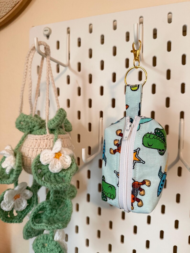May include: A light blue zippered pouch with a cartoon dinosaur print, attached to a gold-colored keychain. A crocheted plant hanger with white flowers hangs nearby. The items are displayed on a white pegboard.