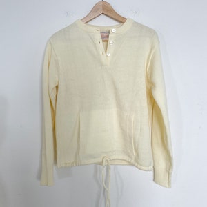 May include: A pale yellow long-sleeved sweater with a buttoned neckline and a drawstring at the bottom. The sweater has a single pocket on the front.