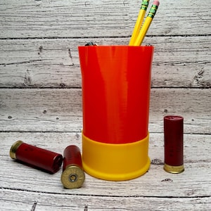 May include: A red and yellow pencil holder resembling a shotgun shell, with three yellow pencils inside. Several red shotgun shells are scattered around the holder. The pencil holder is a unique desk accessory.