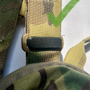 May include: A close-up of a camouflage-patterned strap with a black plastic buckle. The strap is made of woven nylon and has a textured surface.
