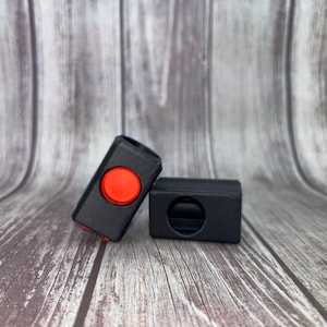 May include: Two black rectangular objects with rounded edges. One object has a red circular button on top. The other object has a circular hole. The objects are on a wooden surface.