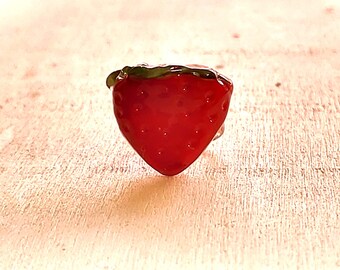 May include: A ring featuring a bright red strawberry charm with a glossy finish and a small green leaf detail at the top. The strawberry has a textured surface with small indentations representing seeds, creating a realistic fruit appearance.