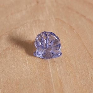 May include: A small, translucent, lavender-colored gemstone carved in the shape of a flower. The gemstone is set against a light brown wooden surface, with visible details in the carving.