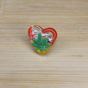 May include: A glass heart-shaped pendant with a green marijuana leaf design. The heart is layered with red, yellow, and clear glass.