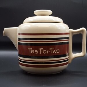 May include: A cream-coloured ceramic teapot with a lid, featuring horizontal stripes in dark brown and black. The text "Tea For Two" is printed in a stylised font across the centre of the body. It includes a side handle and a short spout.