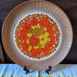 May include: A round serving tray with a white centre featuring a dense, vibrant floral pattern in shades of orange, yellow, and gold. The wide, textured border has a light brown, woven-look finish, and the tray rests on a black stand.