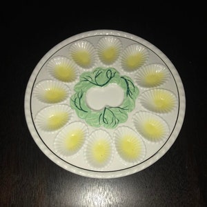 Pode incluir: Um prato de ovos de cerâmica branca com 12 reentrâncias individuais em forma de ovo. O prato tem um desenho floral verde e branco no centro.