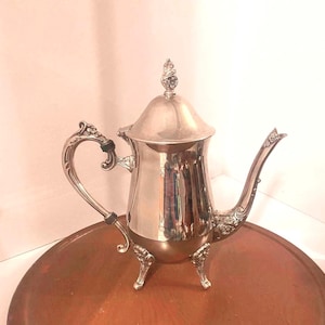 May include: A polished silver-toned metal coffee pot featuring a curved handle with dark accents, a long slender spout, and a domed lid topped with a decorative finial. The vessel stands on three ornate, curved legs with detailed floral motifs.