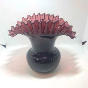 Pode incluir: Um vaso de vidro roxo profundo com uma base bulbosa e uma abertura larga e alargada com uma borda distintamente franzida e ondulada. O material translúcido capta a luz, destacando o corpo liso e arredondado e a borda sinuosa.