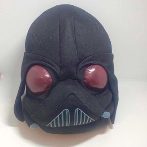 May include: A plush toy shaped like a black helmet with a rounded top, featuring two large, glossy, dark red circular eye lenses, a central triangular nose section, and a gray-lined mouth grill with small light blue accents on the lower sides.