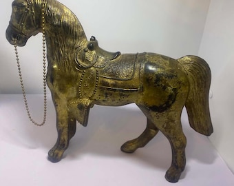 May include: A gold-toned horse figurine featuring a distressed finish, a detailed saddle with embossed patterns, and a thin metallic ball chain acting as a bridle. The horse stands in a profile position with its head turned slightly forward.