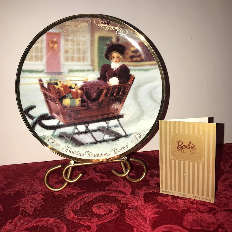 Może przedstawiać: Ozdobny talerz przedstawiający lalkę Barbie w saniach, część kolekcji Holiday Traditions Barbie. Talerz ma złoty brzeg i jest eksponowany na złotym metalowym stojaku. Projekt talerza zawiera zimową scenę.