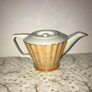 Puede incluir: Una tetera blanca con un borde dorado y un patrón floral marrón. La tetera tiene una tapa y un pico.