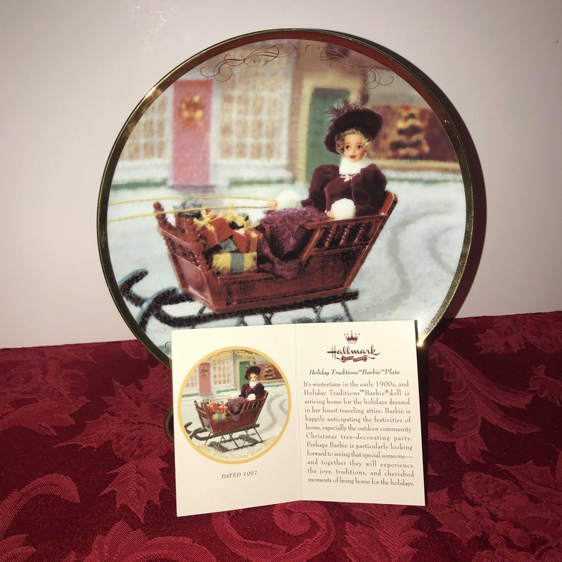 Może przedstawiać: Ozdobny talerz ze złotą obw&oacute;dką, przedstawiający lalkę Barbie w saniach na tle zimowej scenerii. Talerz jest częścią kolekcji Hallmark Holiday Traditions Barbie Plate. Widoczny jest tekst "Holiday Traditions Barbie Plate".